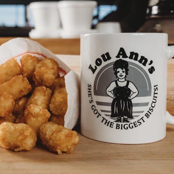 Lou Ann's branded mug next to a paper bag filled with golden-brown tater tots on a wooden surface.
